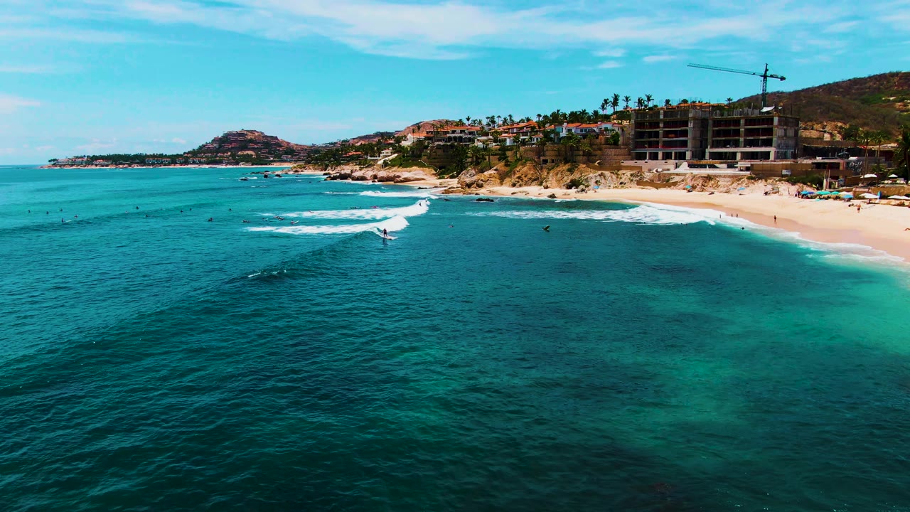 Aerial frame at Punta Buzzards, Baja California Sur showing turquoise ocean in the foreground, a white wave starting to break near the sandy beach, and small people visible in the water and onshore.