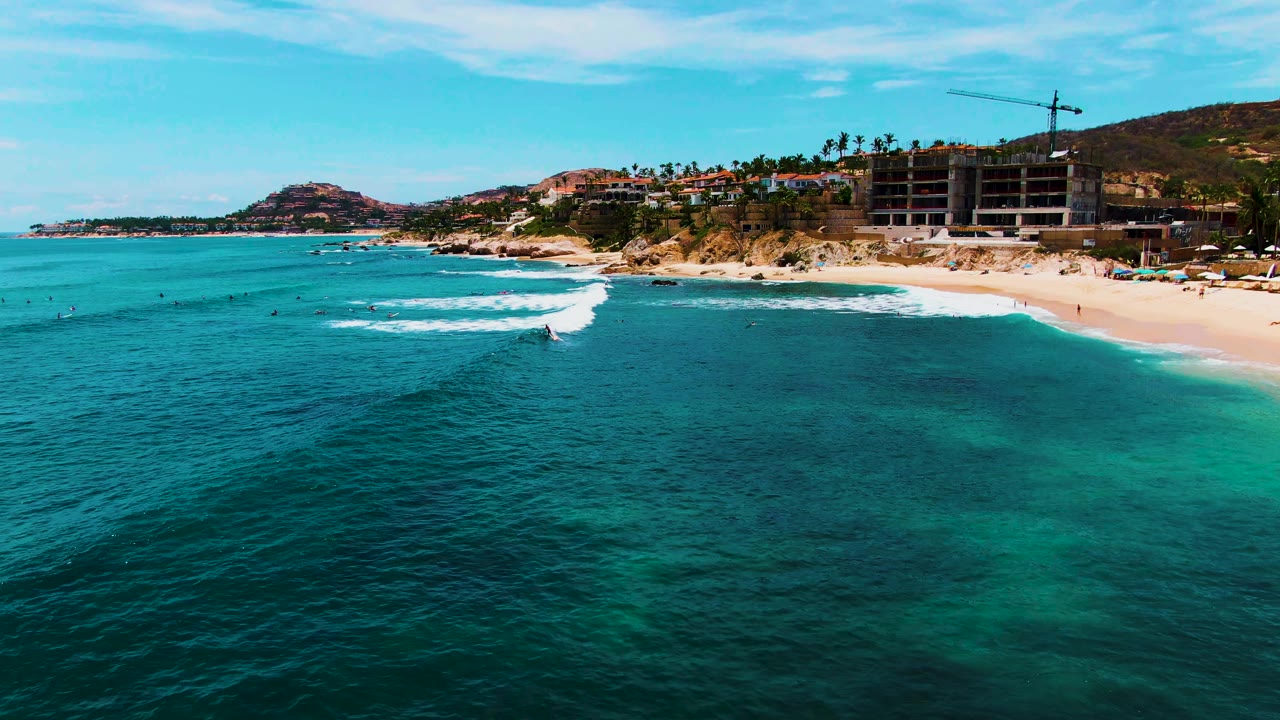 Wide aerial beach view with blue water, white surf, and buildings behind the sand.