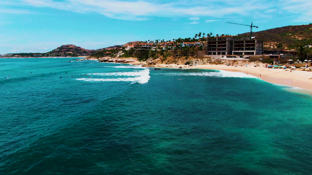 Aerial frame at Punta Buzzards showing darker reef-like patches under clear water, a white breaking wave near the beach, small people in the surf, and low coastal hills behind the shoreline.