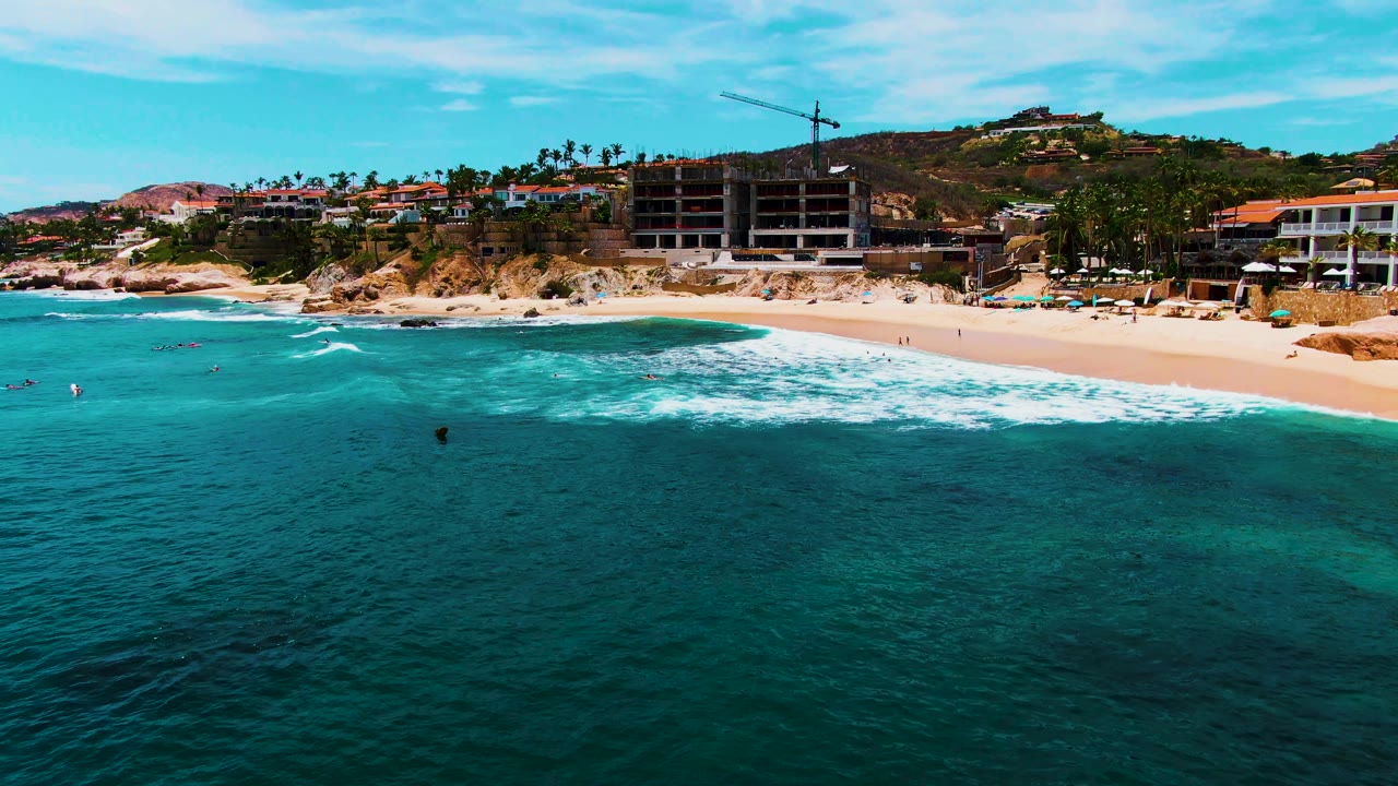 Aerial view of Punta Buzzards in Baja California Sur with turquoise water in the foreground, waves reaching the sand, small swimmers offshore, and hillside buildings behind the beach.