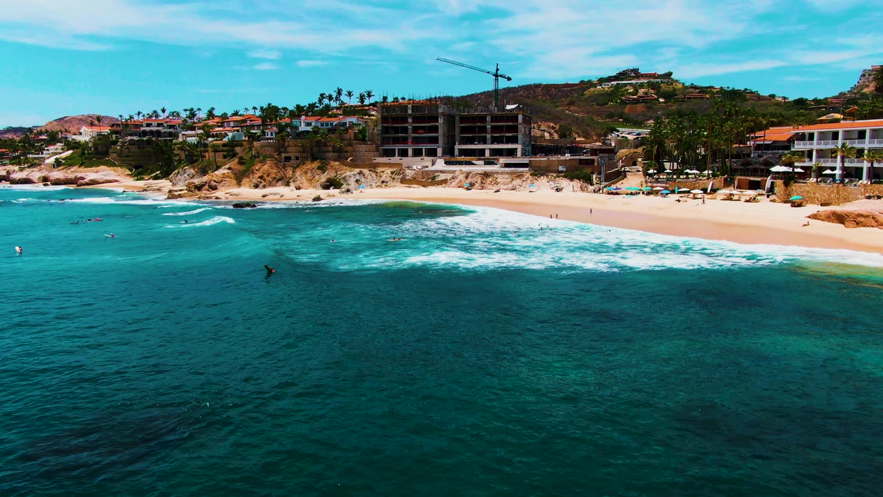 Wide aerial beach view with breaking waves, swimmers in the water, palm trees, buildings, and a crane on the hillside.