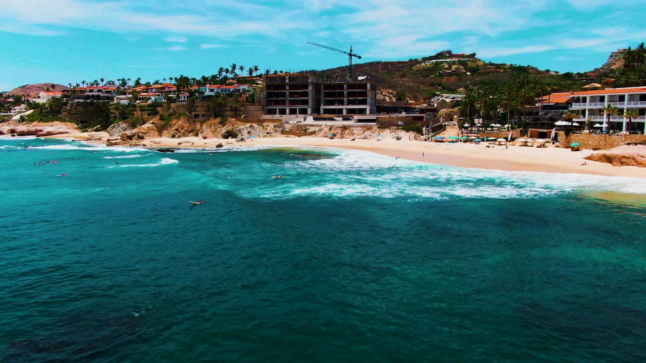 Aerial frame of Buzzards Beach with a broad band of turquoise water, white surf curling toward shore, visible swimmers, beach umbrellas, palms, hillside buildings, and clear sky.