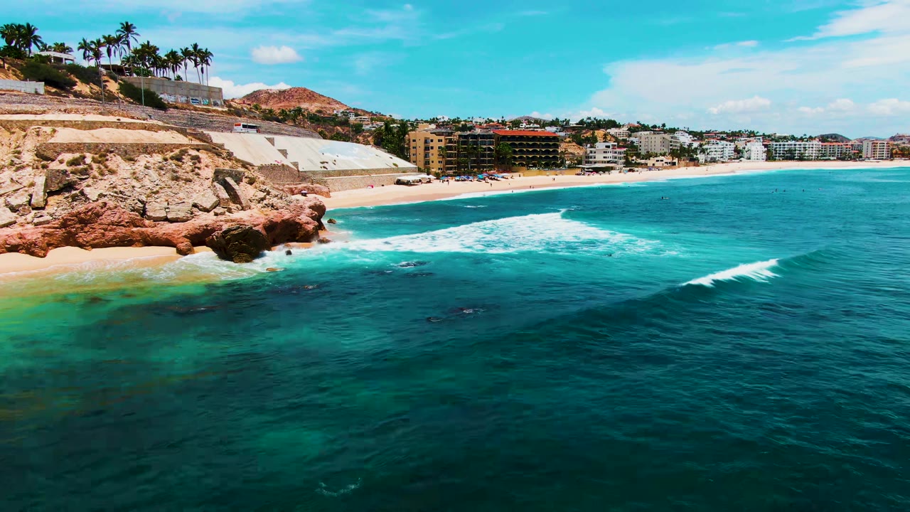 Opening frame of Buzzards Beach in Baja California Sur, with turquoise water in the foreground, a rocky outcrop at left, sandy shore and distant beachfront buildings, and no clearly visible people.