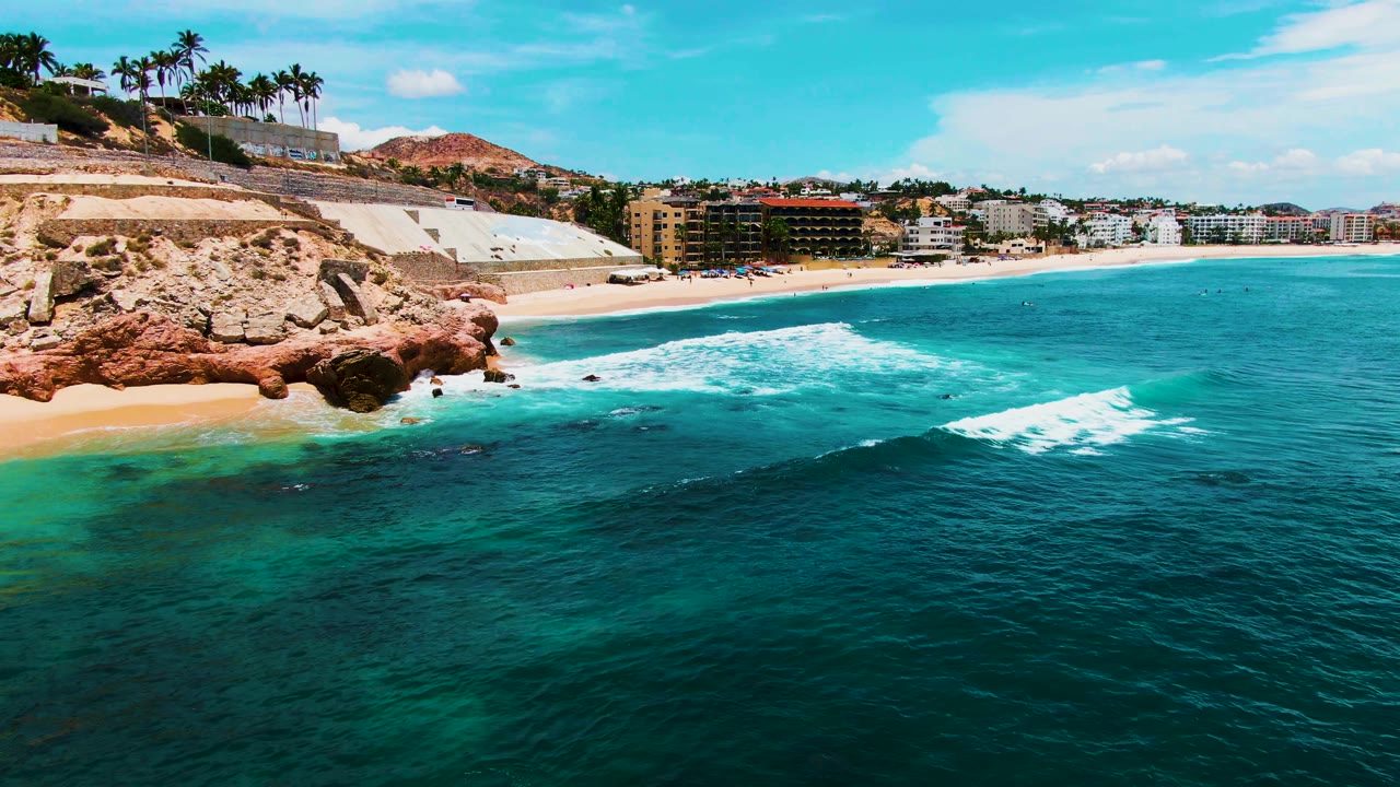 At 0:02, white foam spreads farther across the shallow turquoise water at Buzzards Beach, with the rocky point left of frame and distant buildings along the sand, and no clearly visible people.