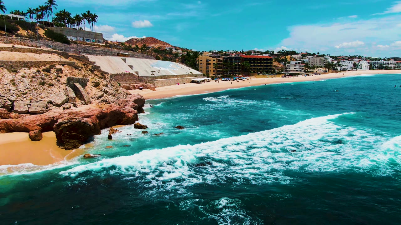 Opening aerial frame at Punta Buzzards shows a foamy wave crossing turquoise water toward a sandy beach beside red rocks, with palm trees and coastal buildings in the background and no visible people.