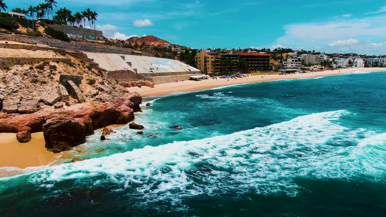 At 0:01, the wave foam widens across the lower frame at Punta Buzzards while the sandy beach, reddish cliff, palm trees, and distant buildings remain visible, with no visible people.