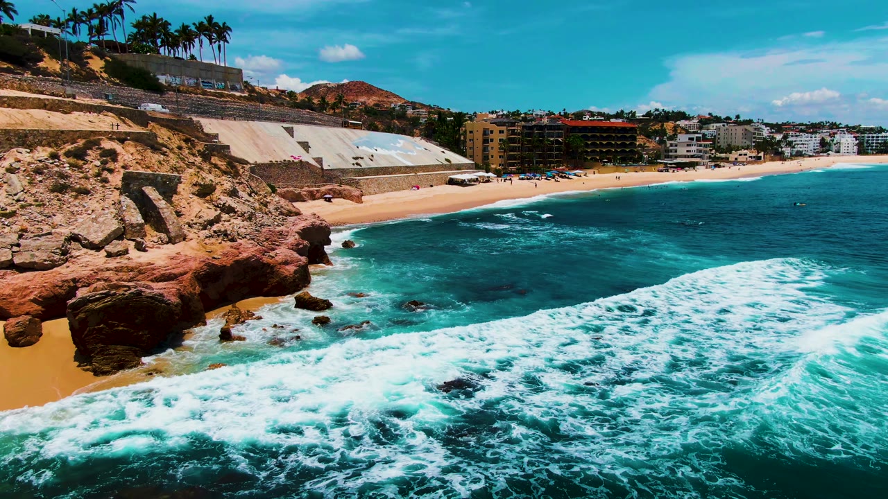 At 0:02, white surf curves diagonally through the foreground at Punta Buzzards, with dark rocks near the beach, a red cliff on the left, and coastal buildings along the shore, with no visible people.