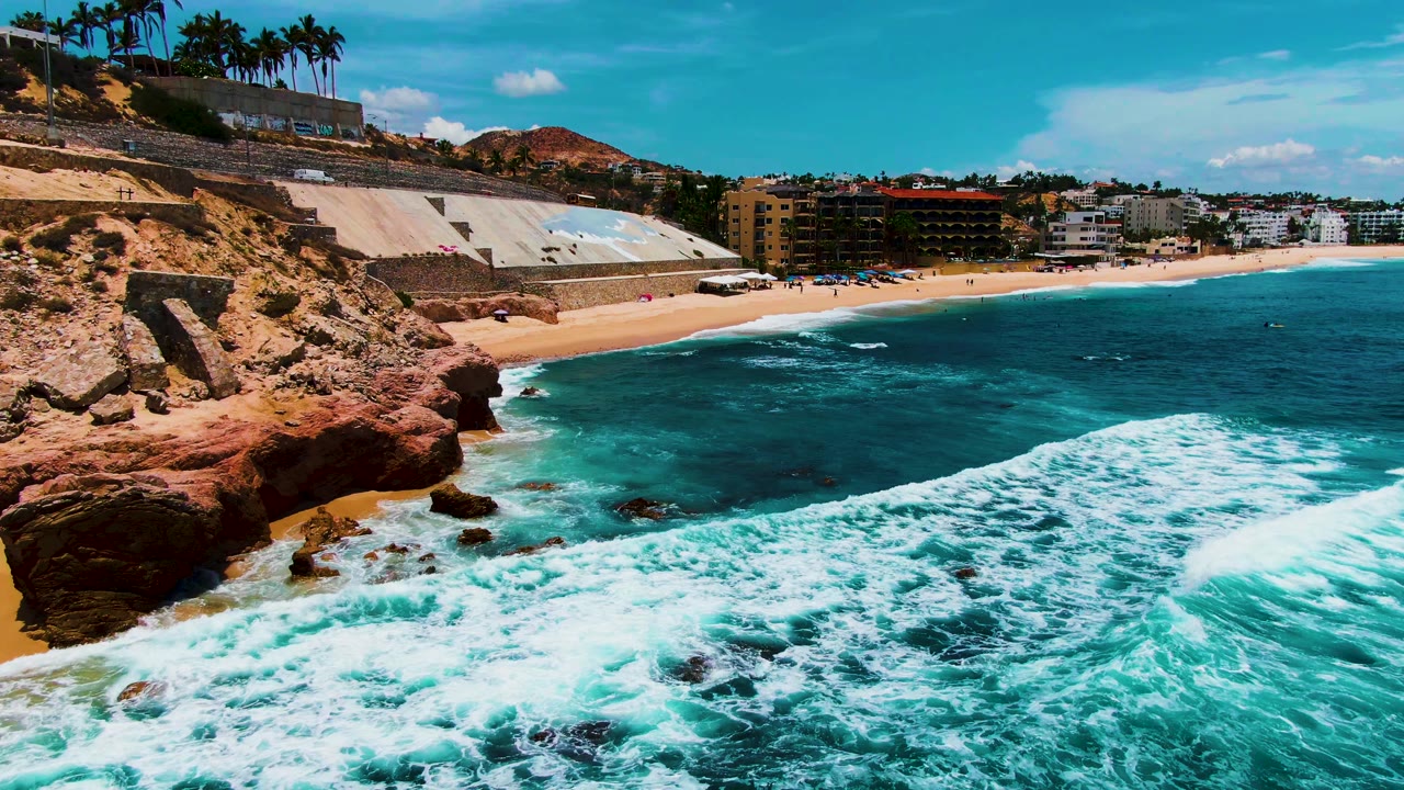 Aerial coastal view with surf foam in the foreground, red rocks, sand, palms, and buildings.