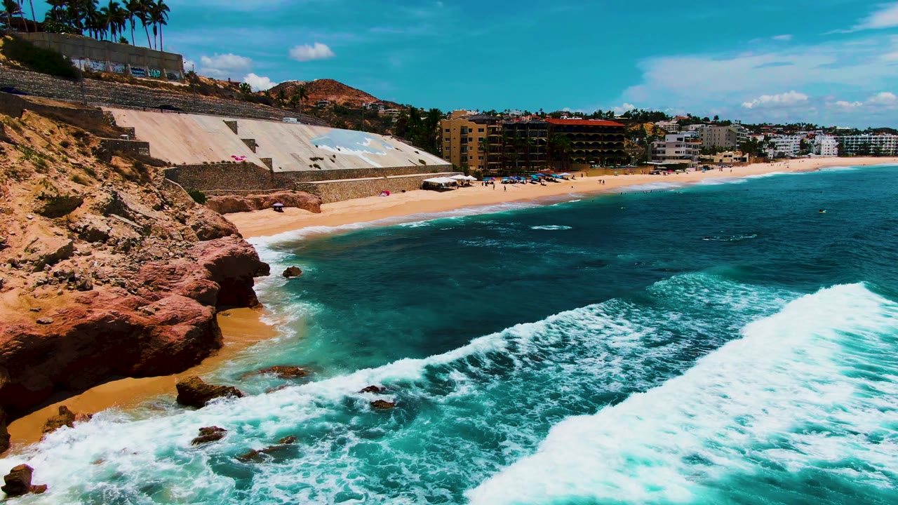 At Buzzards Beach in Baja California Sur, white surf pulls away from the sand beside red shoreline rocks, with a seawall, low buildings, palm trees, and no visible people.