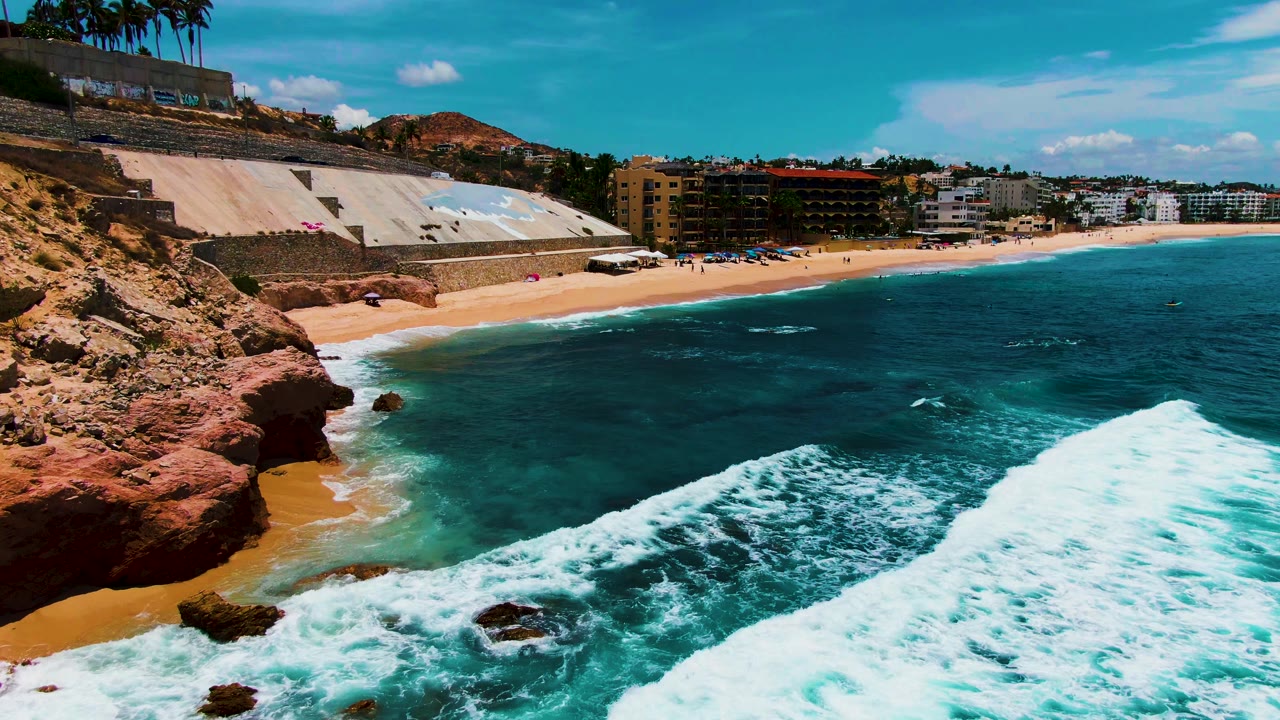 Foamy wave edge recedes along a sandy cove below a seawall and beach buildings.