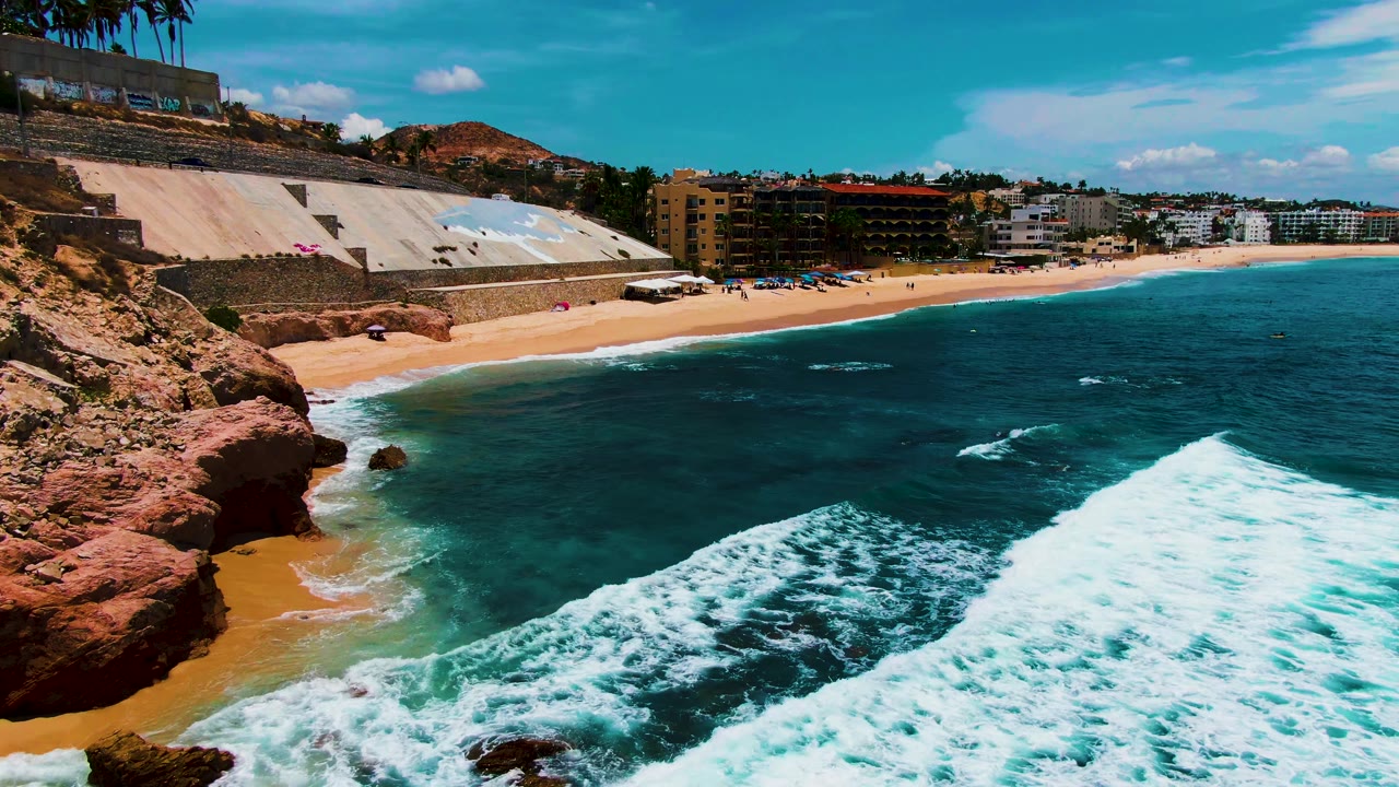 At Buzzards Beach in Baja California Sur, a broad sheet of white foam crosses the lower frame while the sandy beach, seawall, hillside, and distant buildings remain in view, with no visible people.