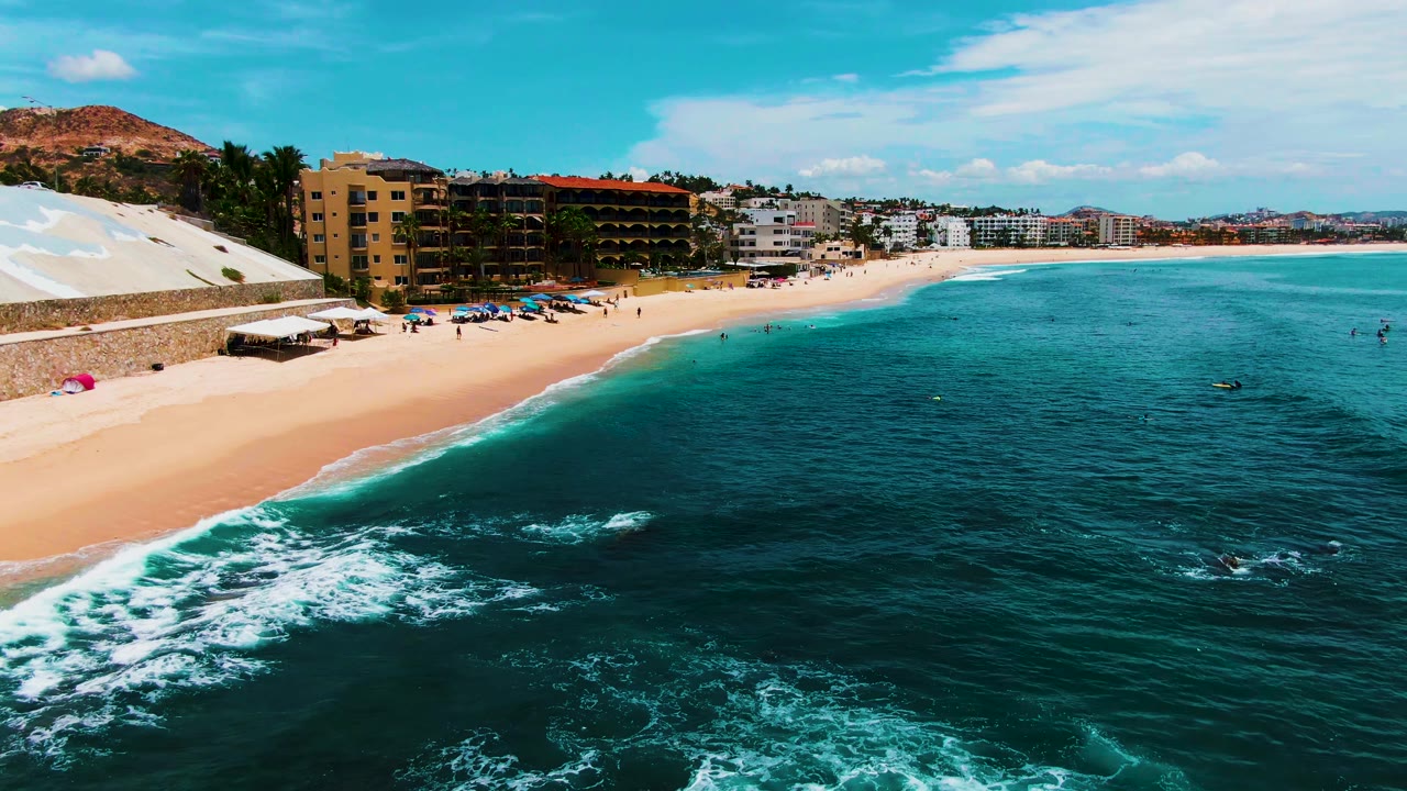 Aerial view of Buzzards Beach in Baja California Sur with turquoise water in the foreground, sandy beach at left, umbrellas, small visible people, and coastal buildings along the shore.