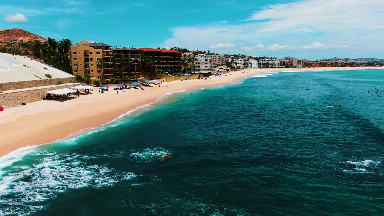 Wide aerial shoreline with beach umbrellas, swimmers, surf, and beachfront buildings.