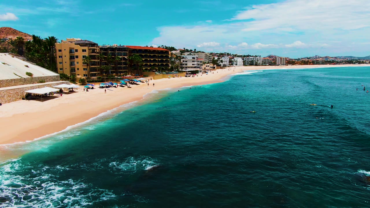 Aerial frame over Buzzards Beach showing darker ocean water in the foreground, pale sand on the left, visible swimmers and beachgoers, umbrellas, and a row of coastal buildings.