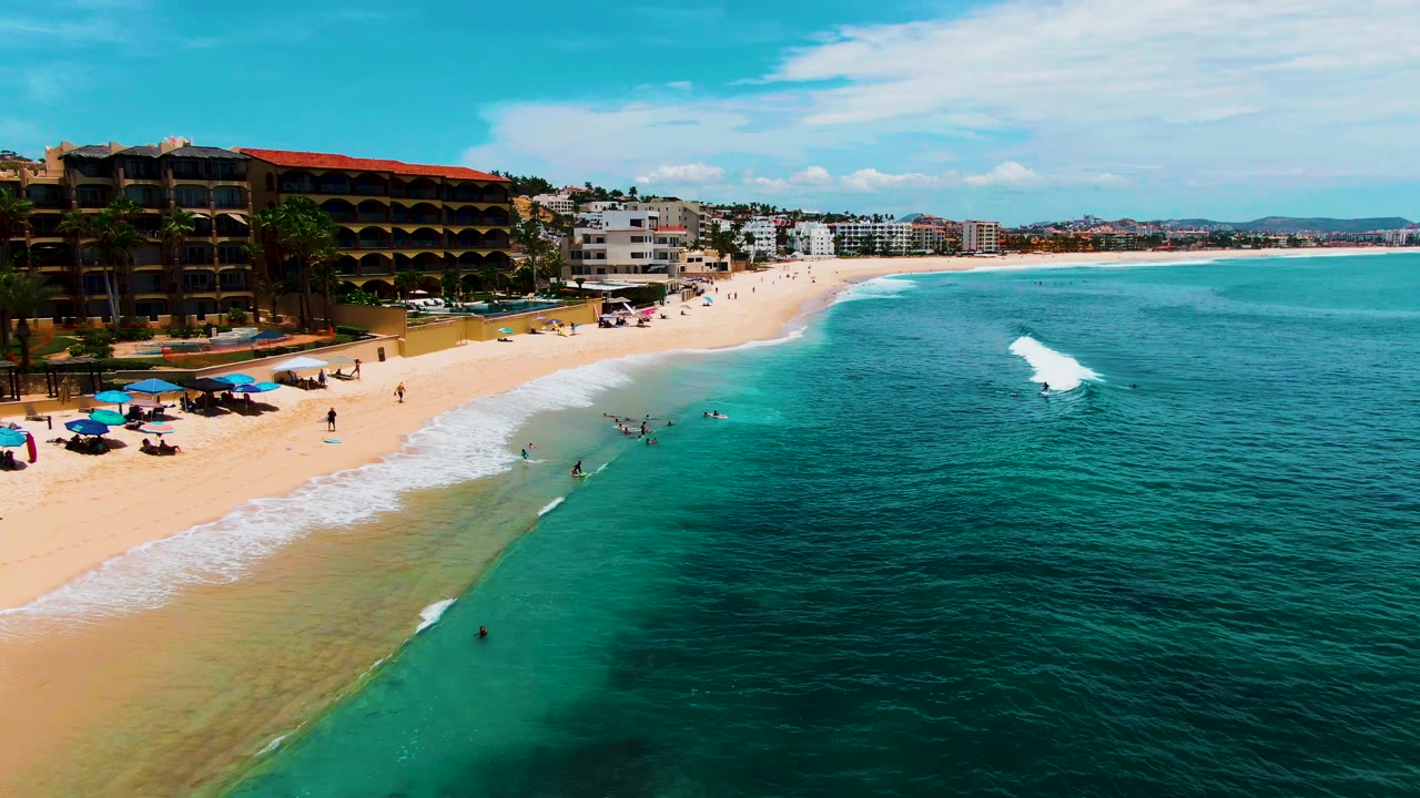Aerial view of Buzzards Beach in Baja California Sur with tan sand on the left, turquoise water on the right, umbrellas near the shore, and small swimmers in the surf.