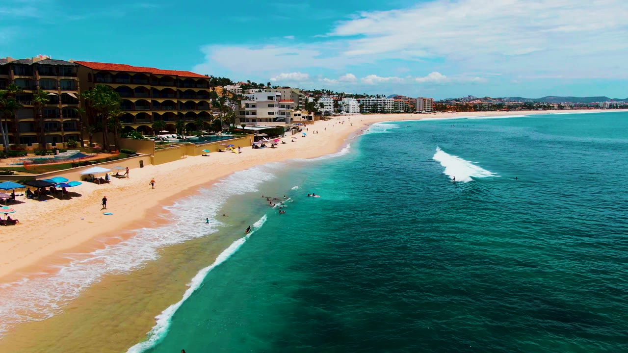 Aerial shoreline view with swimmers beside a forming wave and buildings along the beach.