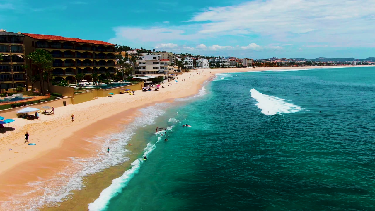 Aerial view of Buzzards Beach as the white wave advances toward swimmers in shallow turquoise water, with sand, umbrellas, beachfront buildings, and distant coast visible.