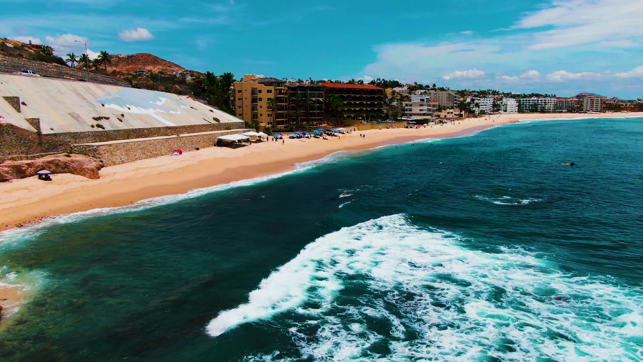 Aerial frame over Buzzards Beach in Baja California Sur, Mexico, with white surf in the foreground, sandy shoreline, small visible beachgoers, and beachfront buildings under a blue sky.