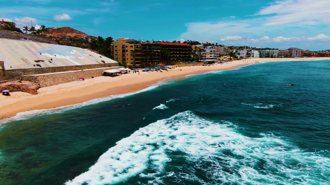White foam spreads across turquoise water beside a sandy beach and coastal buildings.