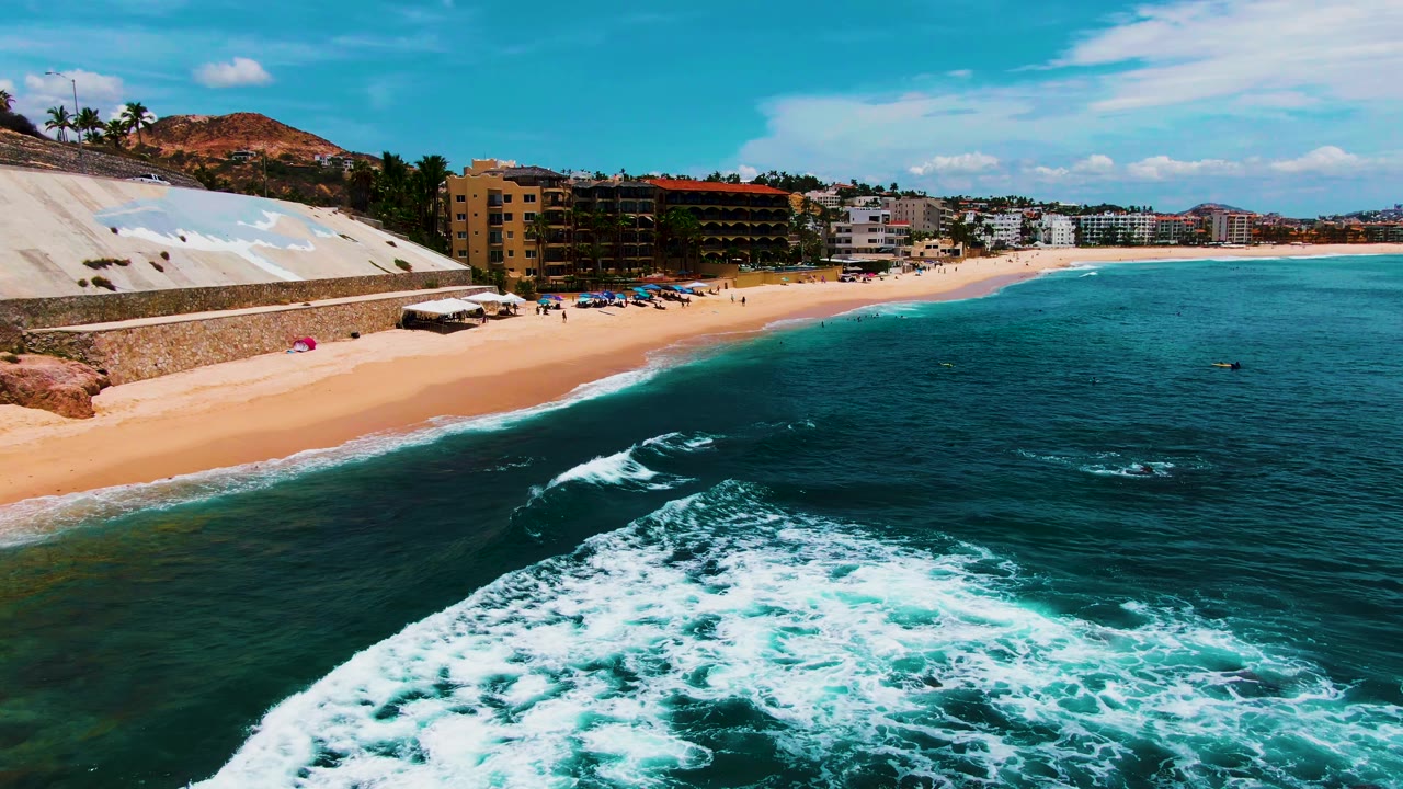 At 0:02, white surf occupies more of the lower frame at Buzzards Beach, while the sandy shoreline, scattered beachgoers, beachfront buildings, hillside, and bright sky remain visible.