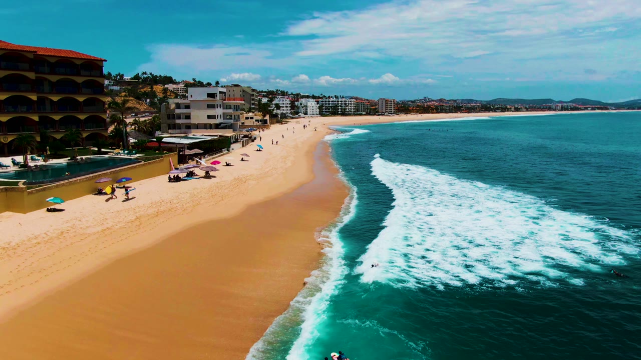 Aerial frame over Buzzards Beach in Baja California Sur with white surf entering from the right, wide sand at center, umbrellas and small beachgoers near the left, and beachfront buildings along the shore.