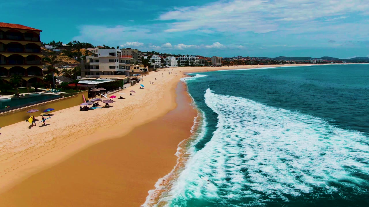 Beach shoreline with waves, umbrellas, people, and low buildings