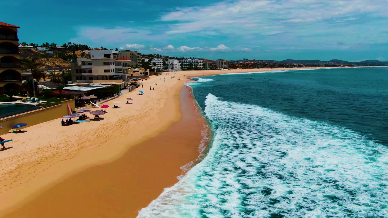 Buzzards Beach appears from an elevated angle with foamy water covering more of the right side, a smooth sand band through the center, small figures on shore, umbrellas at left, and beachfront structures beyond.