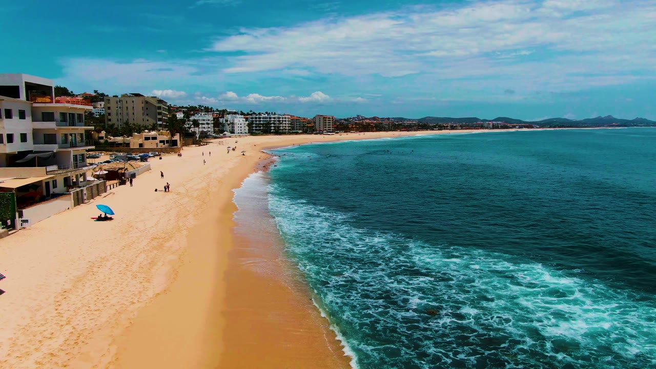 Aerial view of Punta Buzzards beach with sandy shore on the left, teal ocean on the right, white foam at the waterline, scattered people, a blue umbrella, and beachfront buildings under a blue sky.