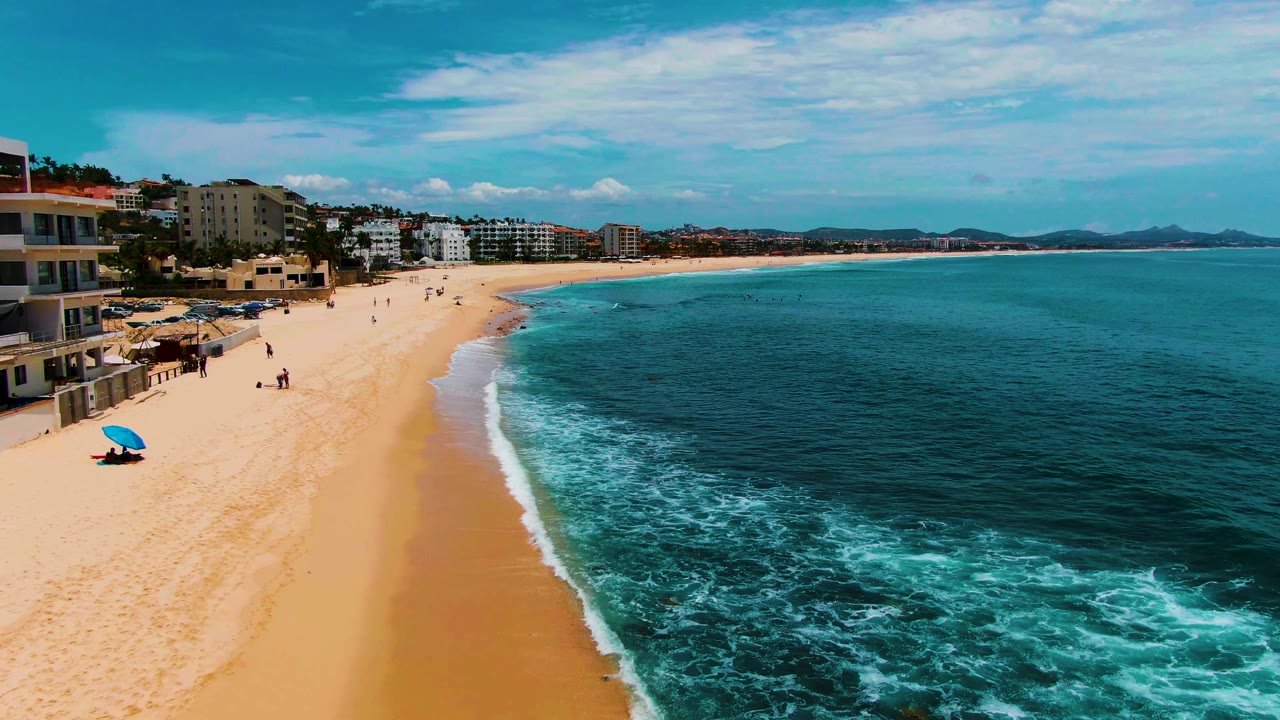 Wide aerial beach view with waves rolling toward sand and people near the shoreline.
