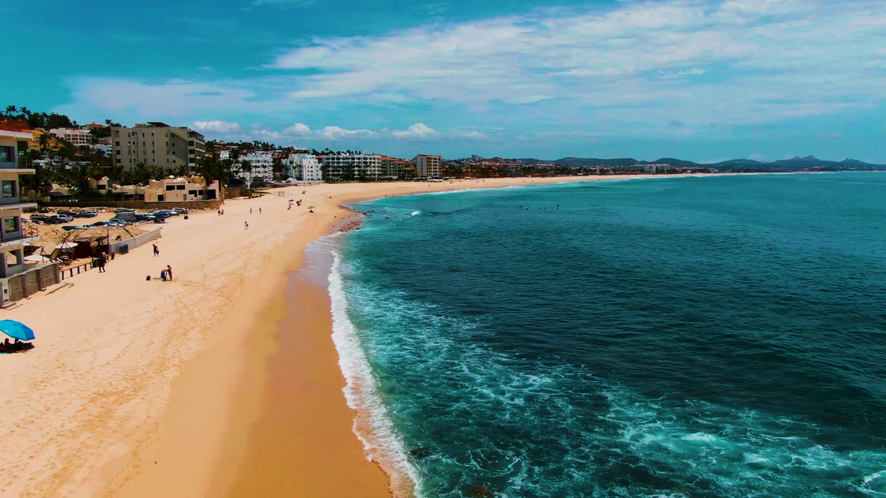 Aerial view of Punta Buzzards beach with shoreline buildings tight to the left edge, open sand with scattered people, white surf foam near the center, and broad teal water stretching to distant hills.