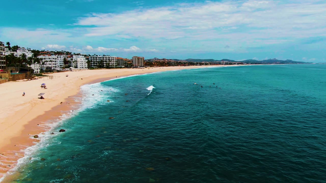Aerial frame over Buzzards Beach in Baja California Sur with pale sand on the left, turquoise water on the right, distant low buildings, and no clearly visible people.