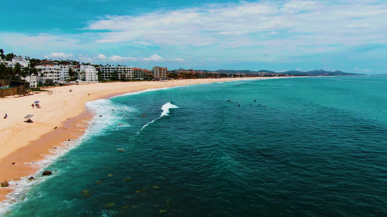 At 0:01, the white wave line sits farther into the nearshore water at Buzzards Beach, with beachfront buildings and distant hills behind the sand and no clearly visible people.