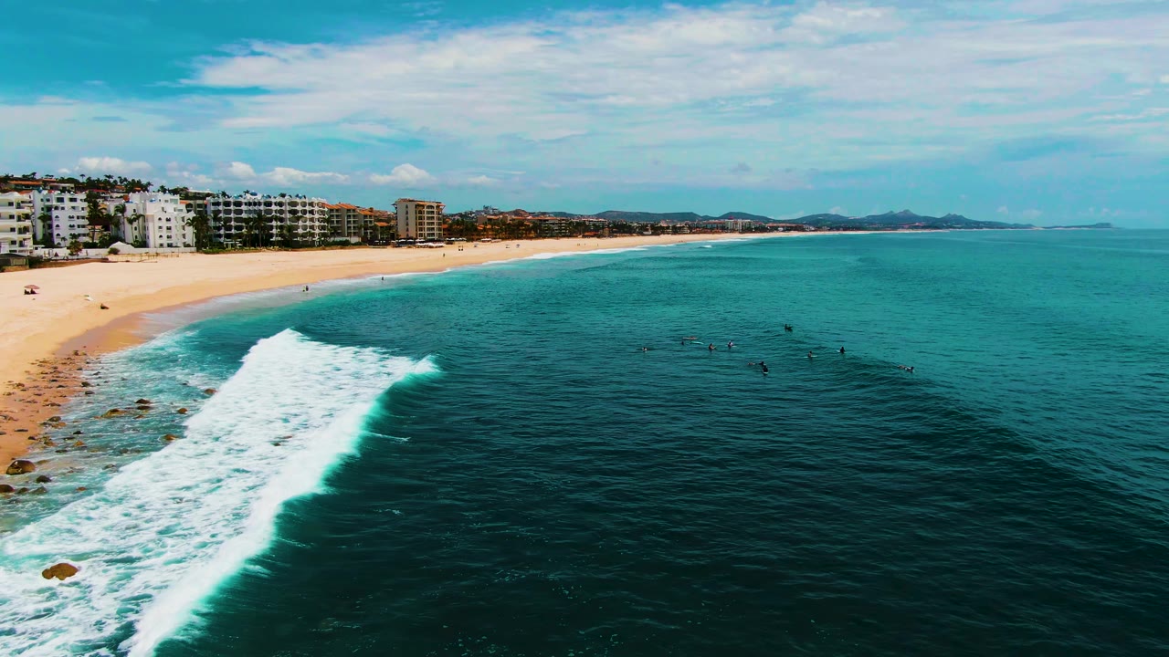 At Buzzards Beach in Baja California Sur, a foamy wave breaks near the sandy shore while small surfers and swimmers are visible in the blue-green water.