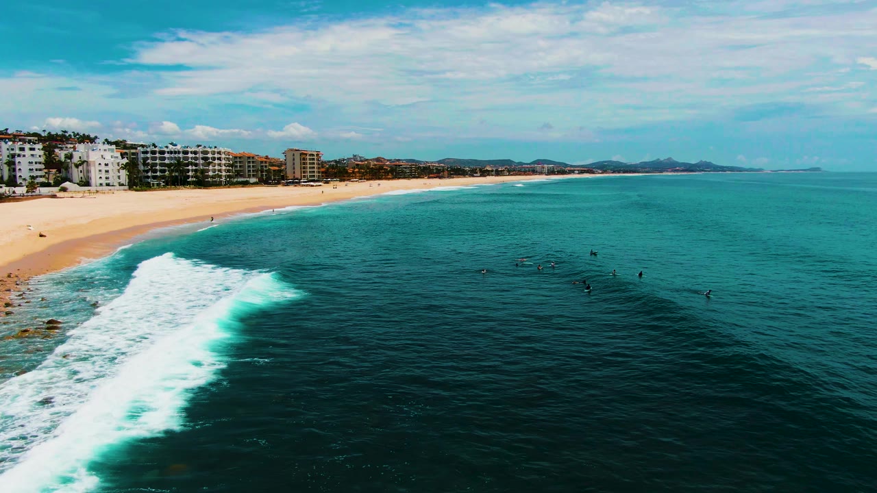 Wide aerial shoreline with wave foam and surfers in the ocean.