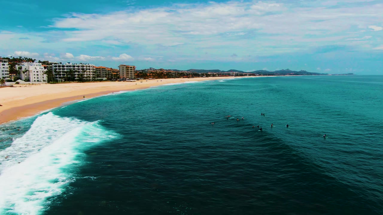 At Buzzards Beach in Baja California Sur, the breaking wave curves from the lower left toward the center while scattered surfers sit beyond the foam line.