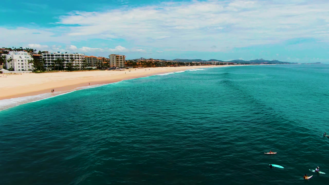 Aerial frame at Punta Buzzards, Baja California Sur showing turquoise ocean, sandy beach, shoreline buildings, and several small people with boards or kayaks near the lower edge.