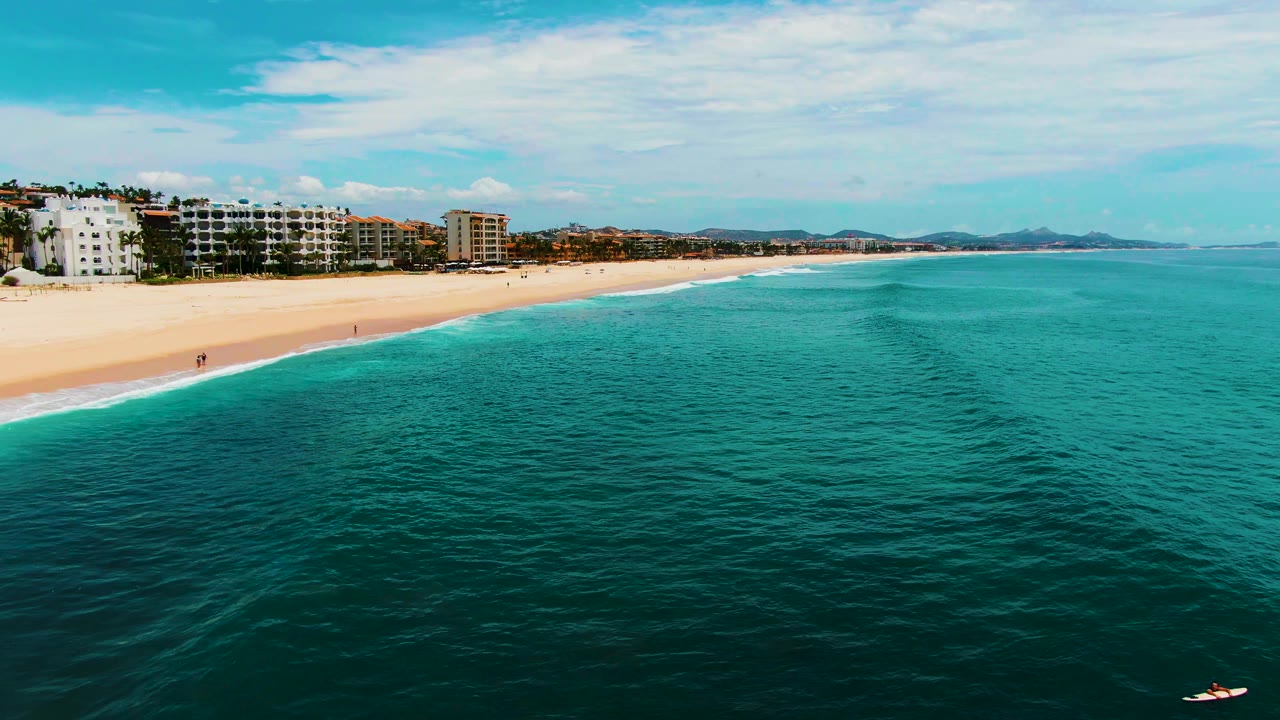 Wide aerial coast view with turquoise water, sandy beach, buildings, and small people near the bottom edge.