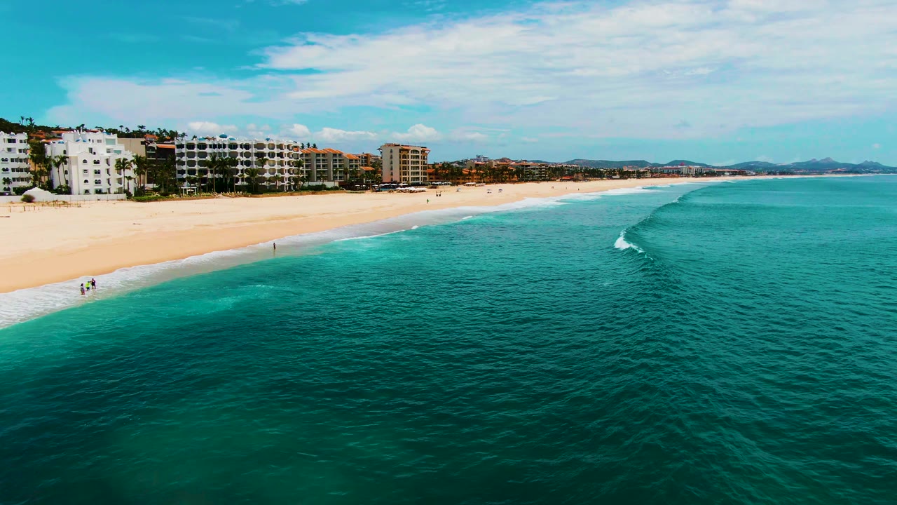 At Punta Buzzards in Baja California Sur, turquoise ocean fills the foreground beside a sandy beach lined with low buildings, with small people faintly visible near the shoreline.