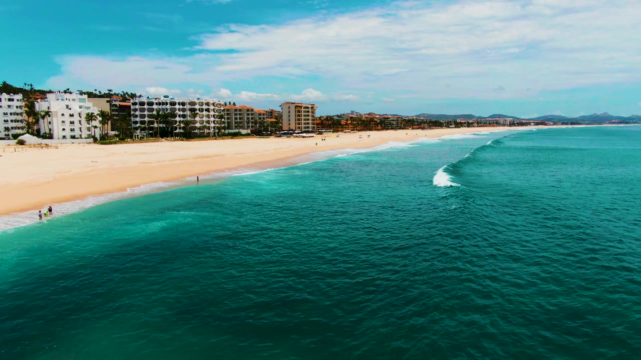 Aerial shoreline view with a small breaking wave offshore and buildings along the beach.