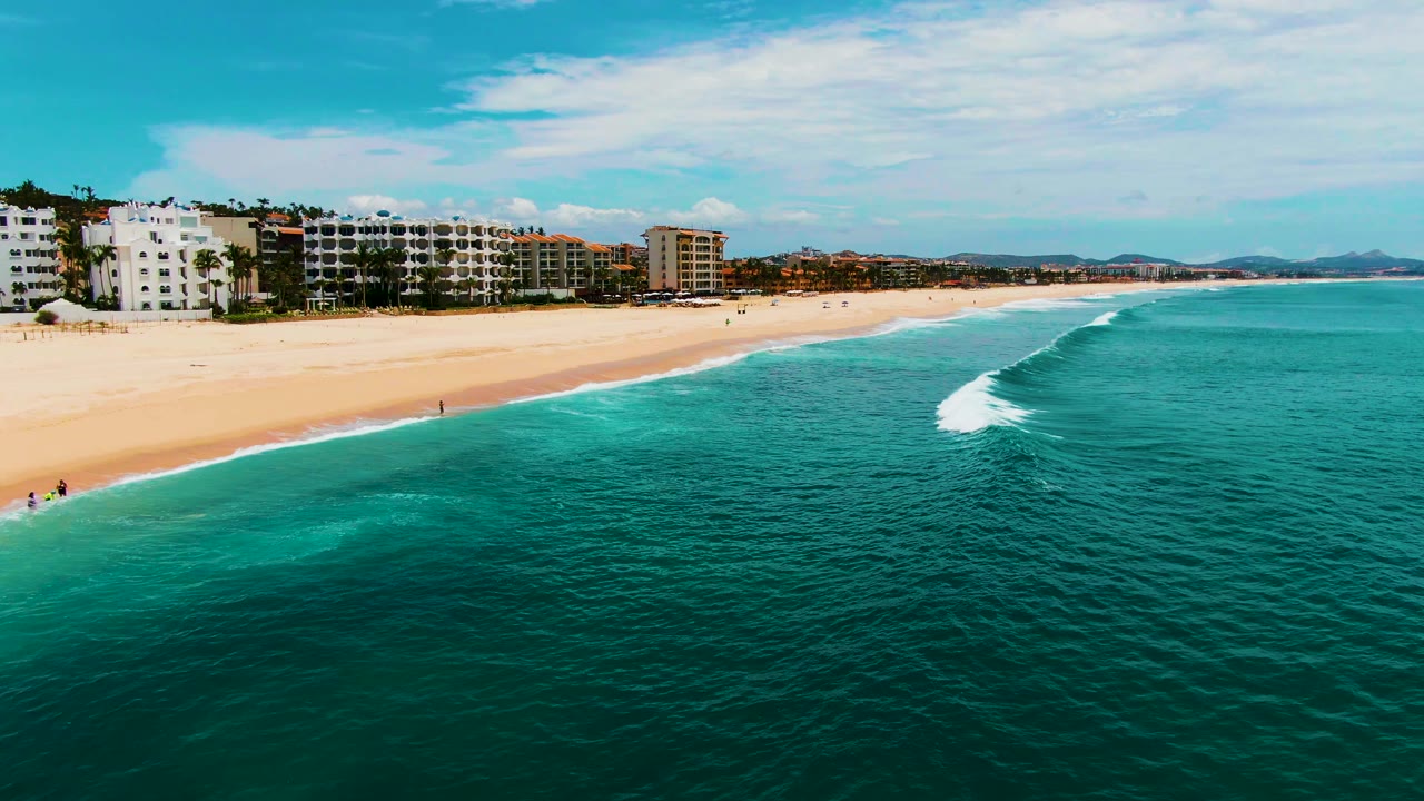 At Punta Buzzards in Baja California Sur, the white surf line has moved closer to the sand, with deep turquoise water in front, beach buildings behind, and small people visible along the shore.