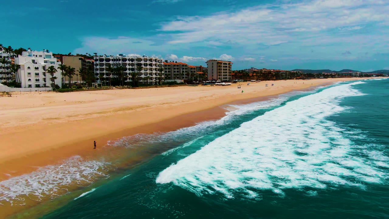 Aerial view at Buzzards Beach in Baja California Sur, Mexico, with turquoise surf entering from the lower right, broad sand at left, coastal buildings in the background, and a small distant person near the waterline.