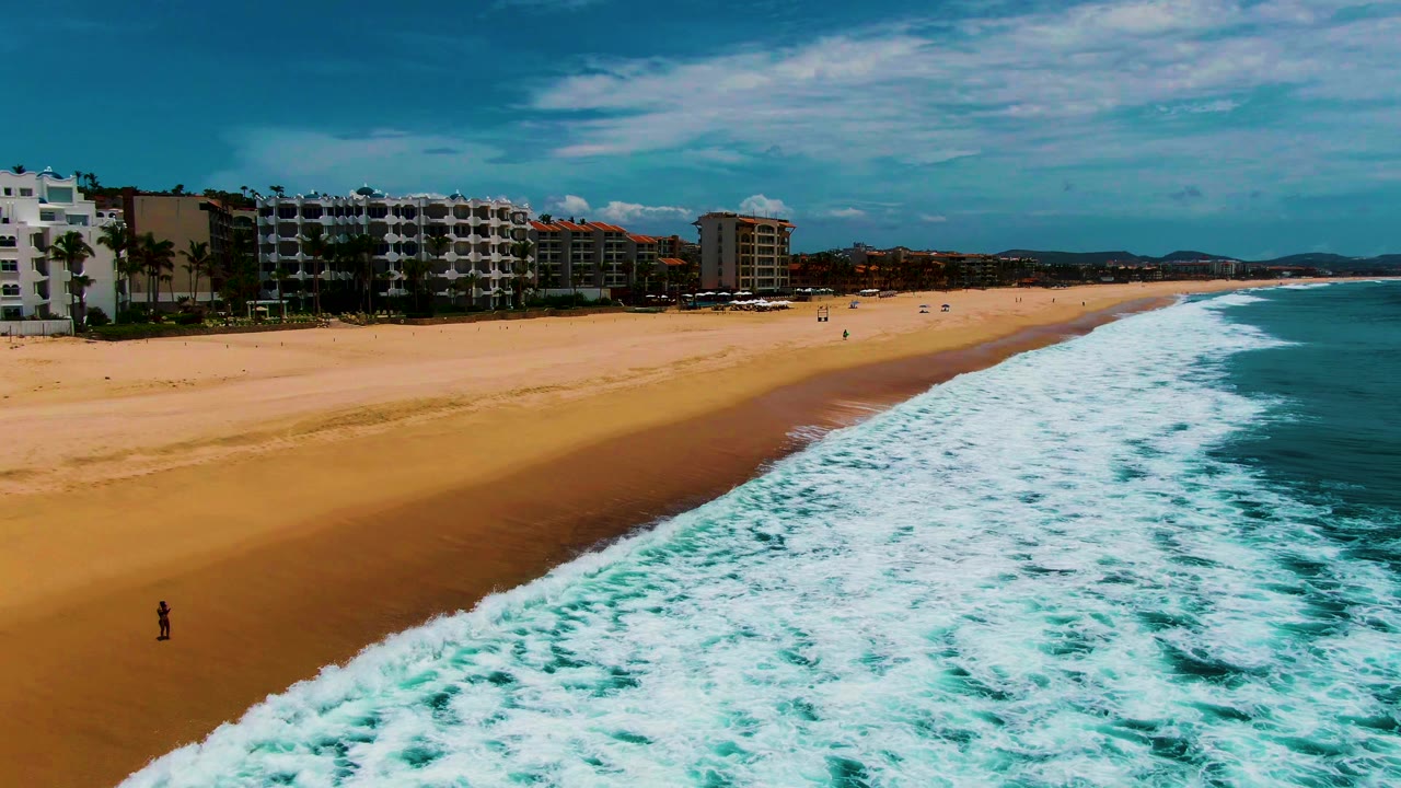 Aerial view over Buzzards Beach as foamy turquoise water covers more of the lower frame, with wide open sand, beachfront buildings, scattered distant beachgoers, and blue sky above.