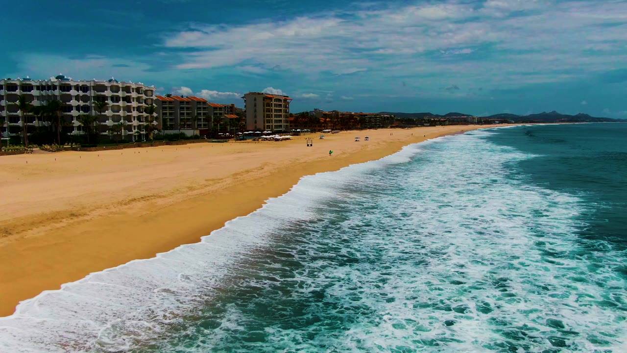 Aerial view at Punta Buzzards, Baja California Sur, with foamy turquoise surf filling the lower right, a wide sandy beach on the left, and coastal buildings along the upper left.