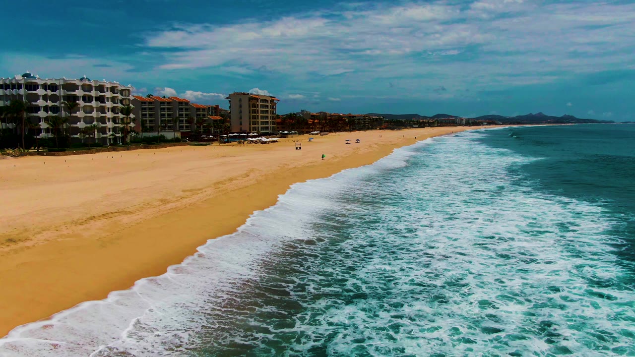 Foamy waves roll onto a sandy beach with buildings set back from the shore.