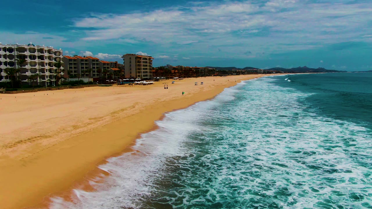 At 0:02, white surf spreads farther across the right side of the frame while the sandy beach and beachfront buildings remain visible along the left side of Punta Buzzards.