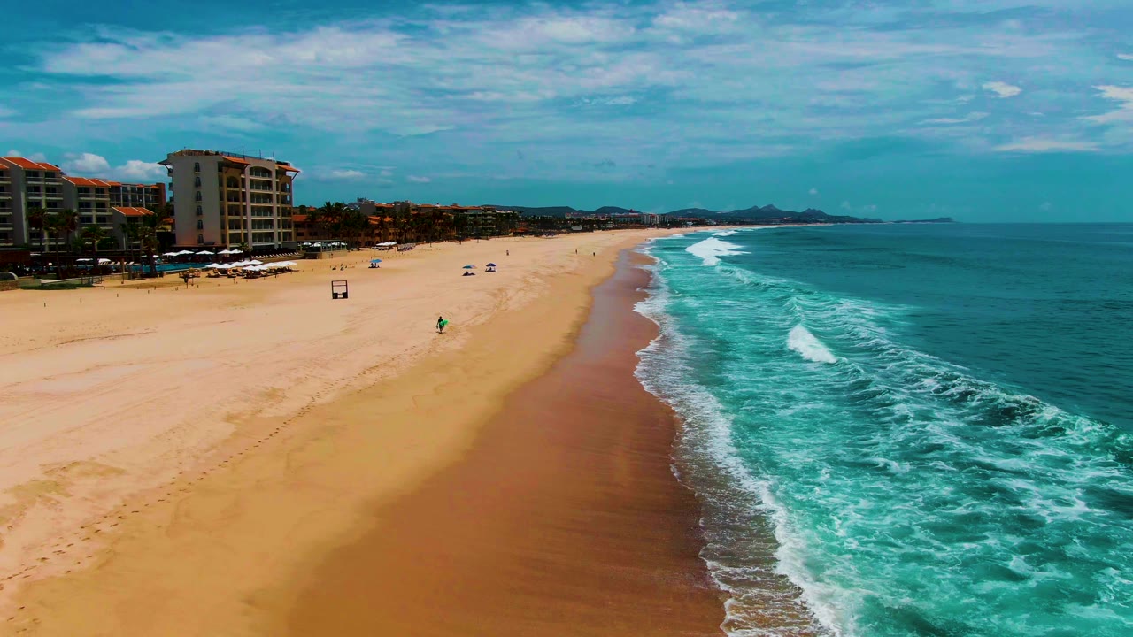 Aerial frame from Punta Buzzards, Baja California Sur, showing a broad sandy beach, turquoise surf, beachfront buildings at left, umbrellas, and small people visible on the sand.