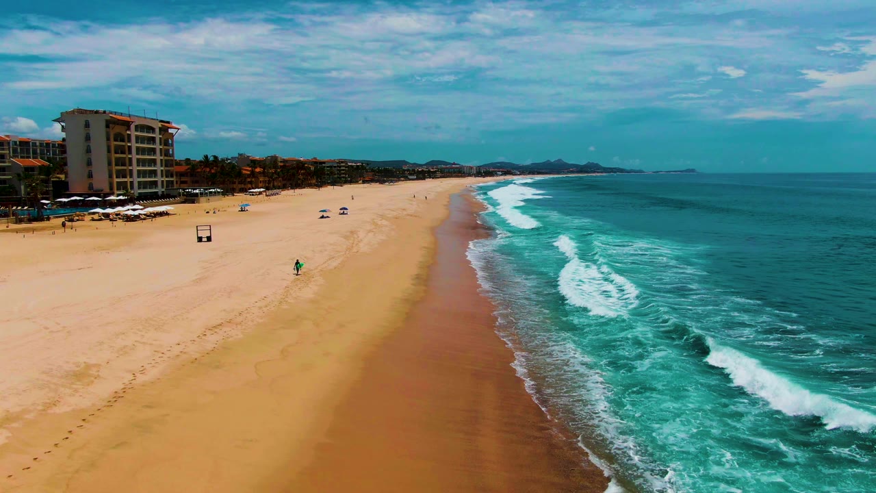 Aerial frame from Punta Buzzards, Baja California Sur, showing a foamy wave band along the right edge, open sand in the center, umbrellas near the upper beach, and buildings along the left background.