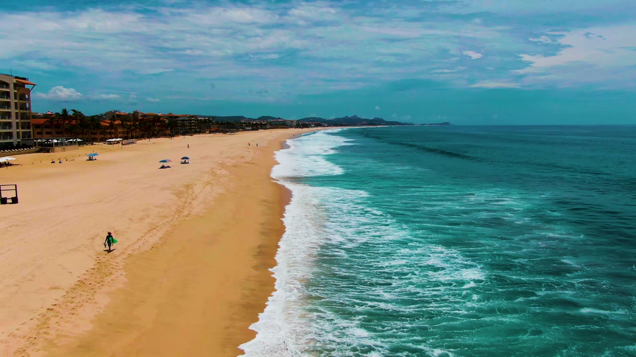 Aerial view at Punta Buzzards showing sandy beach on the left, turquoise ocean on the right, white surf, umbrellas, and a distant person carrying a surfboard.