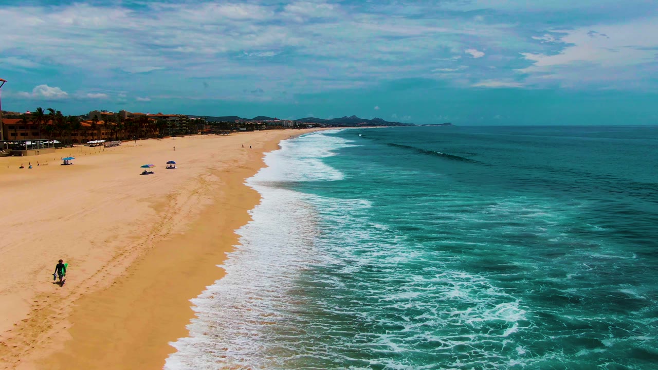 Beach aerial with a person carrying a surfboard near the lower left and waves along shore.