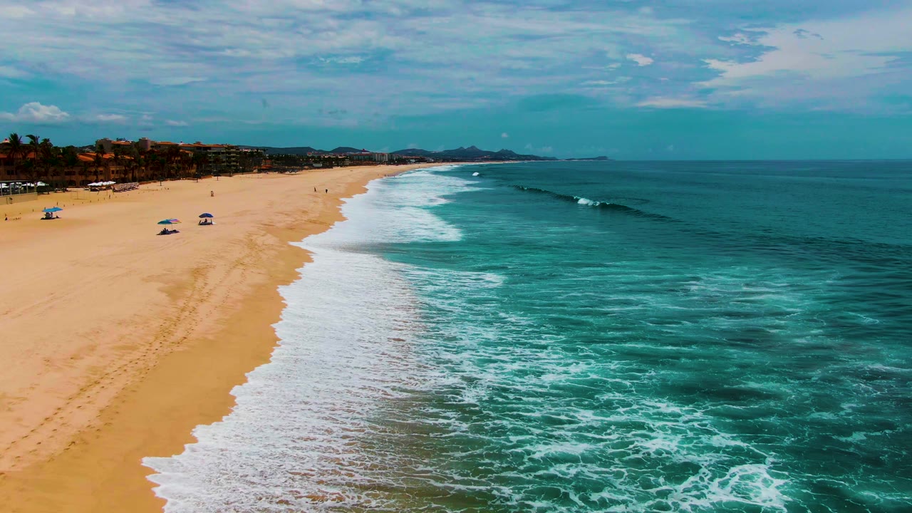 A wider Punta Buzzards shoreline view shows umbrellas on the sand, foamy surf curving along the beach, turquoise water, distant buildings, and no clearly visible people.